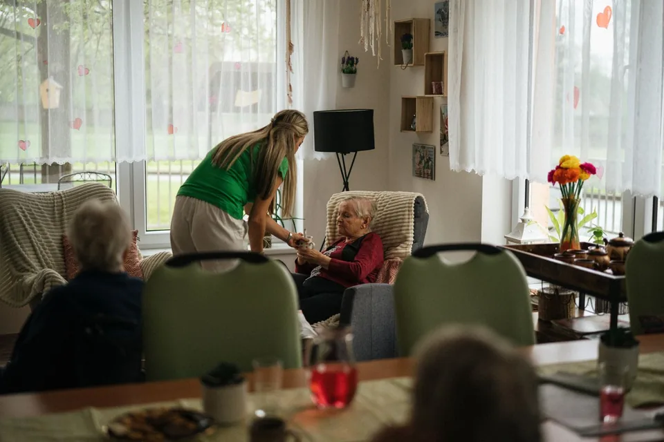 An adult handing a mug to an older woman in a communal room.